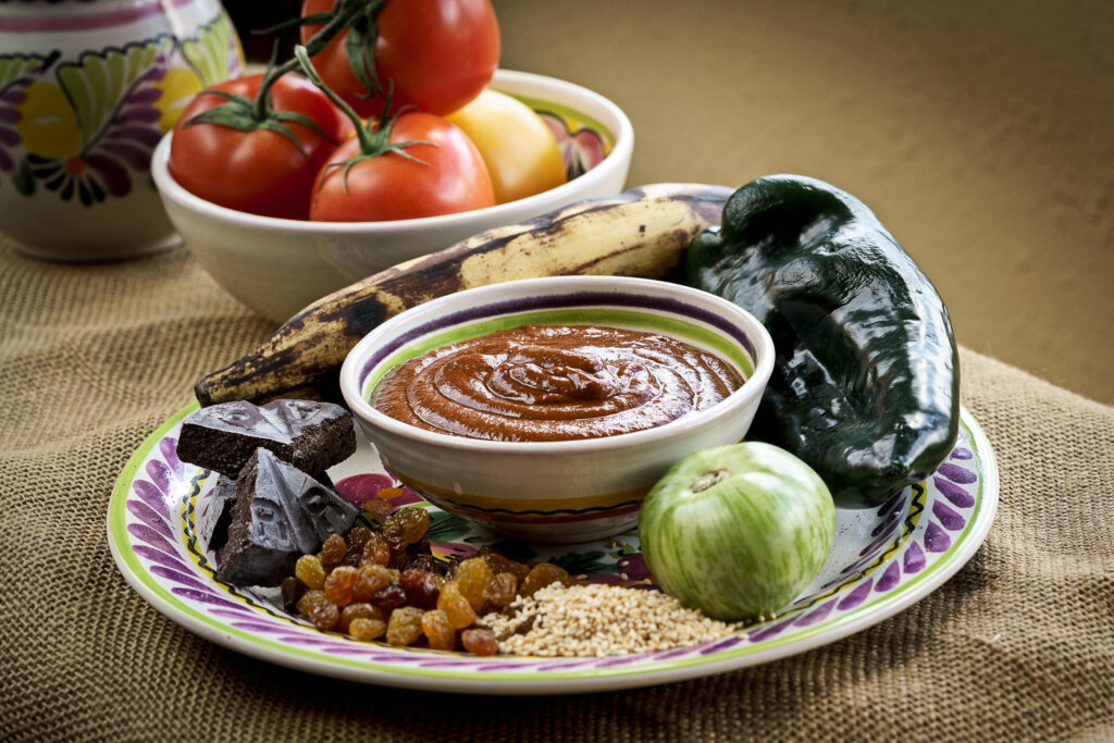 Still-life of a colorful plate with chocolate pieces, dried fruit, and nuts, plus a bowl of mole sauce and fresh vegetables in the background (tomatoes and peppers).