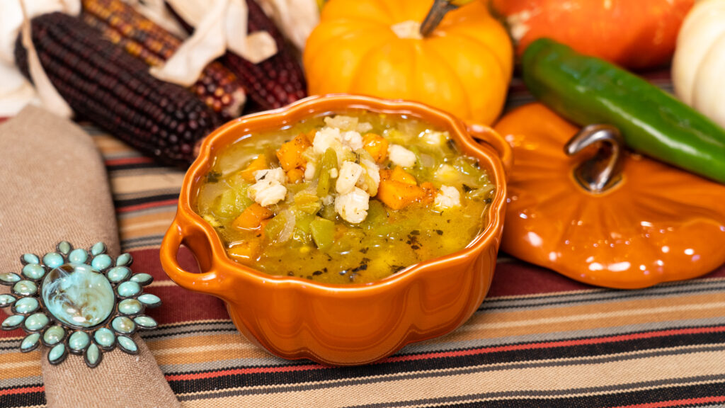 Orange ceramic bowl of vegetable soup with squash and herbs, topped with crumbles, on a striped tablecloth with pumpkins and peppers nearby.