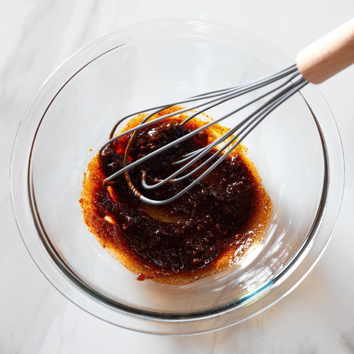 Whisk stirring a dark brown sauce in a clear glass bowl on a light countertop