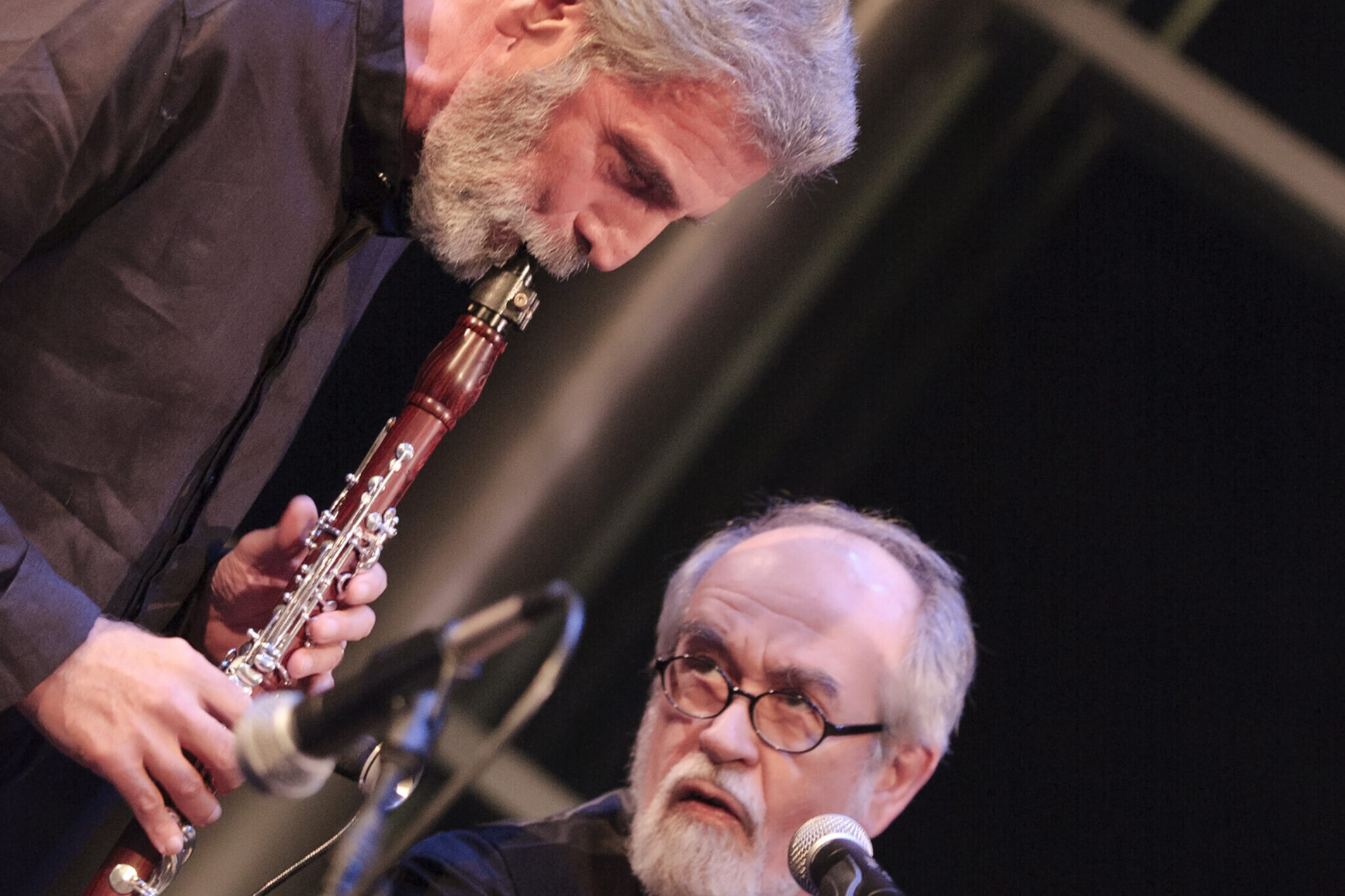 Two older men on stage performing; one plays a clarinet while the other speaks into a microphone, both focused on the music.