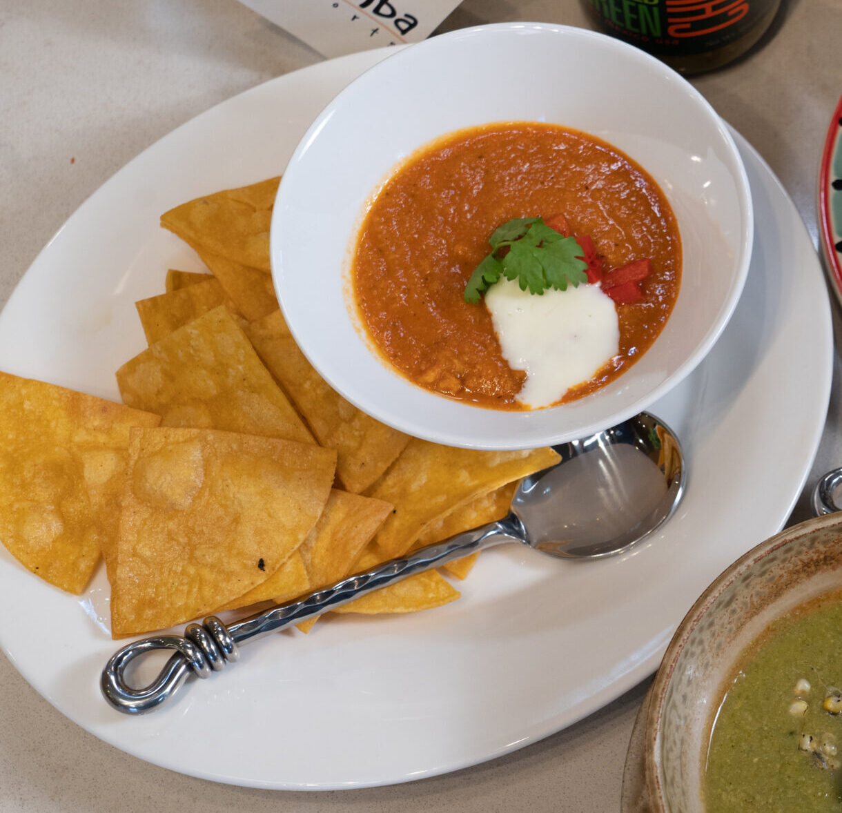 Plate of tortilla chips with red salsa, a dollop of sour cream, and cilantro on top; next to bowls of green soup and a cilantro-lime soup in a colorful setting.