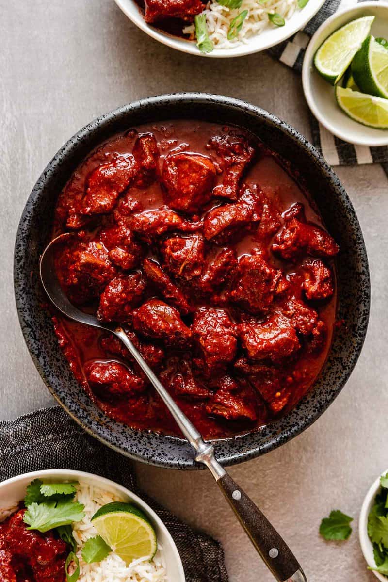 Bowl of red beef stew with chunky meat in thick sauce and a spoon resting inside, surrounded by bowls with lime and rice.