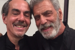 Eddi and Lee smiling and taking a close-up selfie, wearing black outfits, posing together indoors.