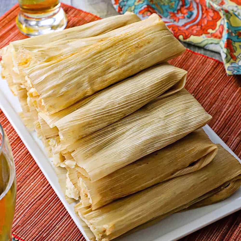 Stack of tamales wrapped in corn husks on a white platter.