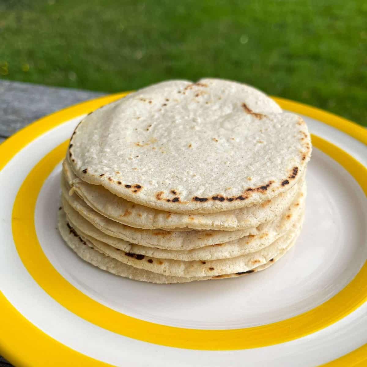 Stack of flour tortillas piled on a white plate with a yellow rim, outdoors on green grass.