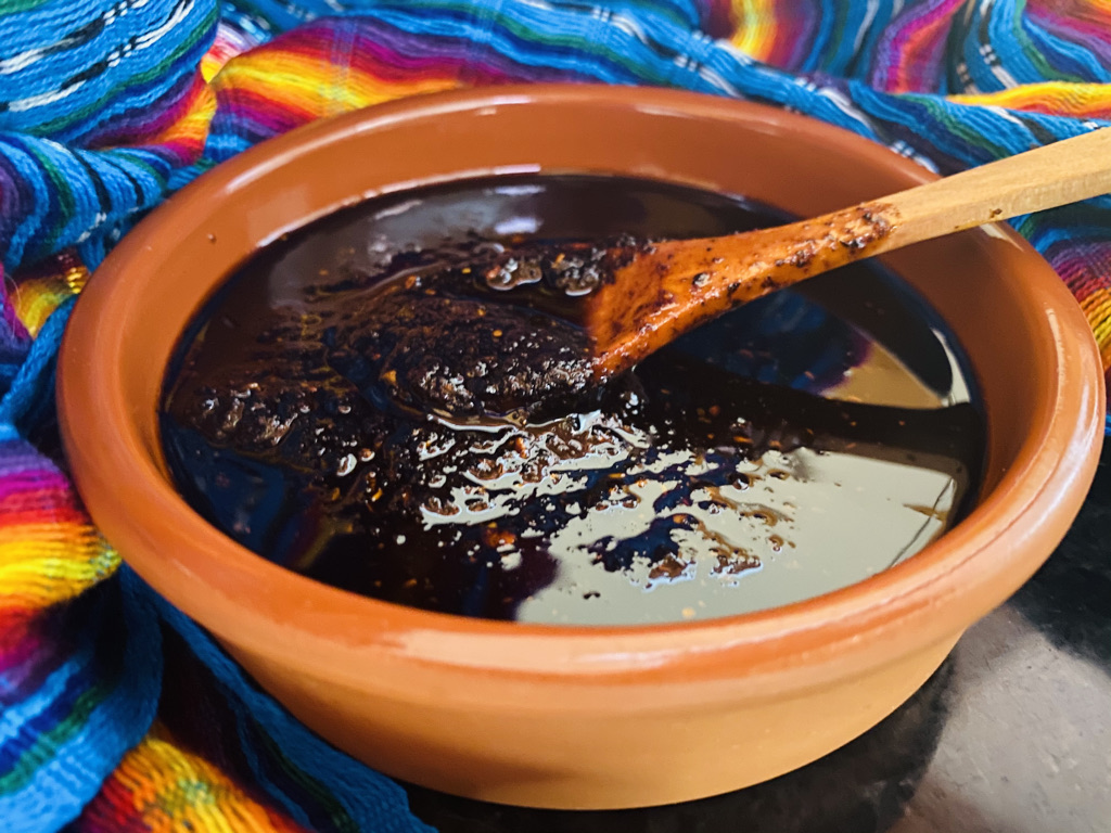Terracotta bowl filled with thick dark chocolate sauce, wooden spoon resting in it, on a vibrant woven textile backdrop.