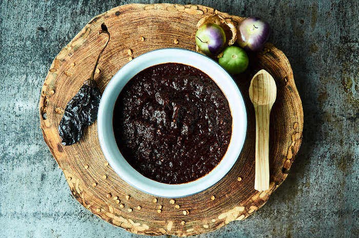 White bowl filled with thick dark paste sits on a rustic wooden board, with a dried chili pepper, small onions, and a wooden spoon nearby.
