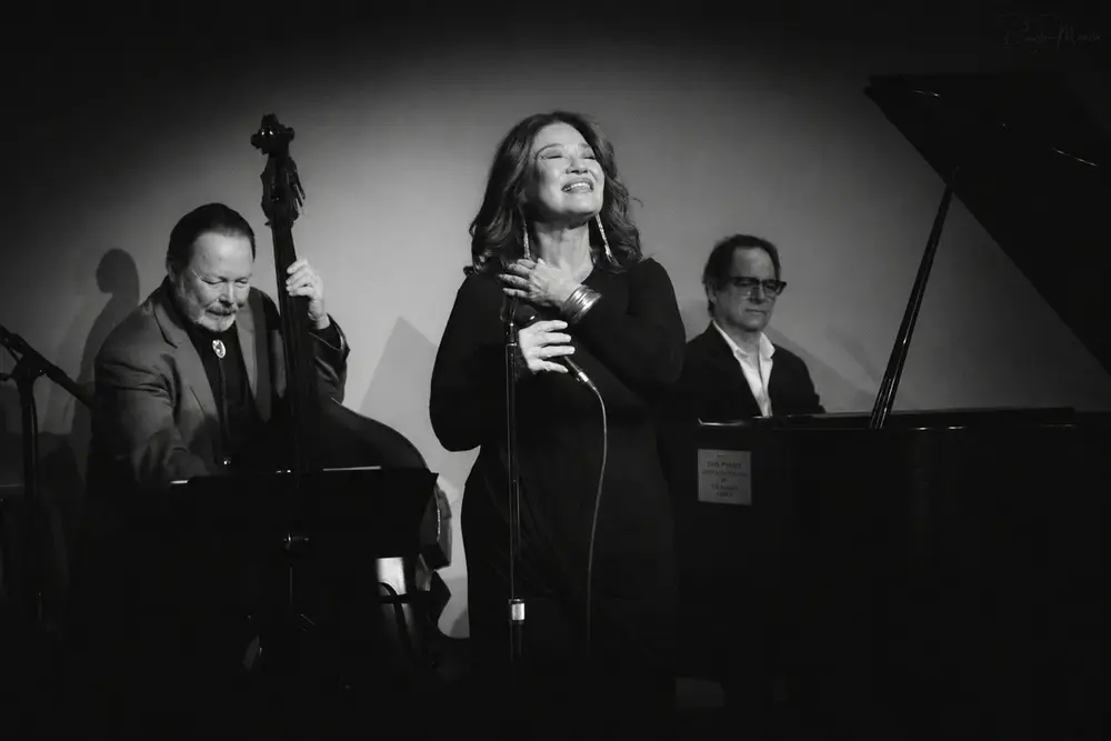 Black-and-white photo of a female jazz singer center stage, with a bassist on the left and a pianist on the right accompanying her on a dimly lit club stage.